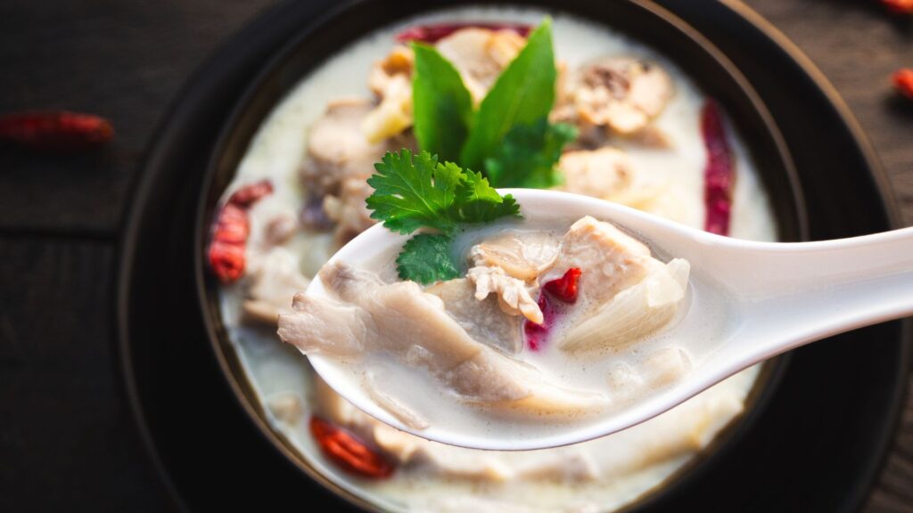 A spoon of creamy Tom Kha Gai soup with chicken, herbs, and chili, served in a bowl on a dark surface.